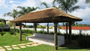 Um quiosque de palha santa fé com um banco branco embaixo e com árvores em sua volta e com um caminho de pedras até o quiosque.